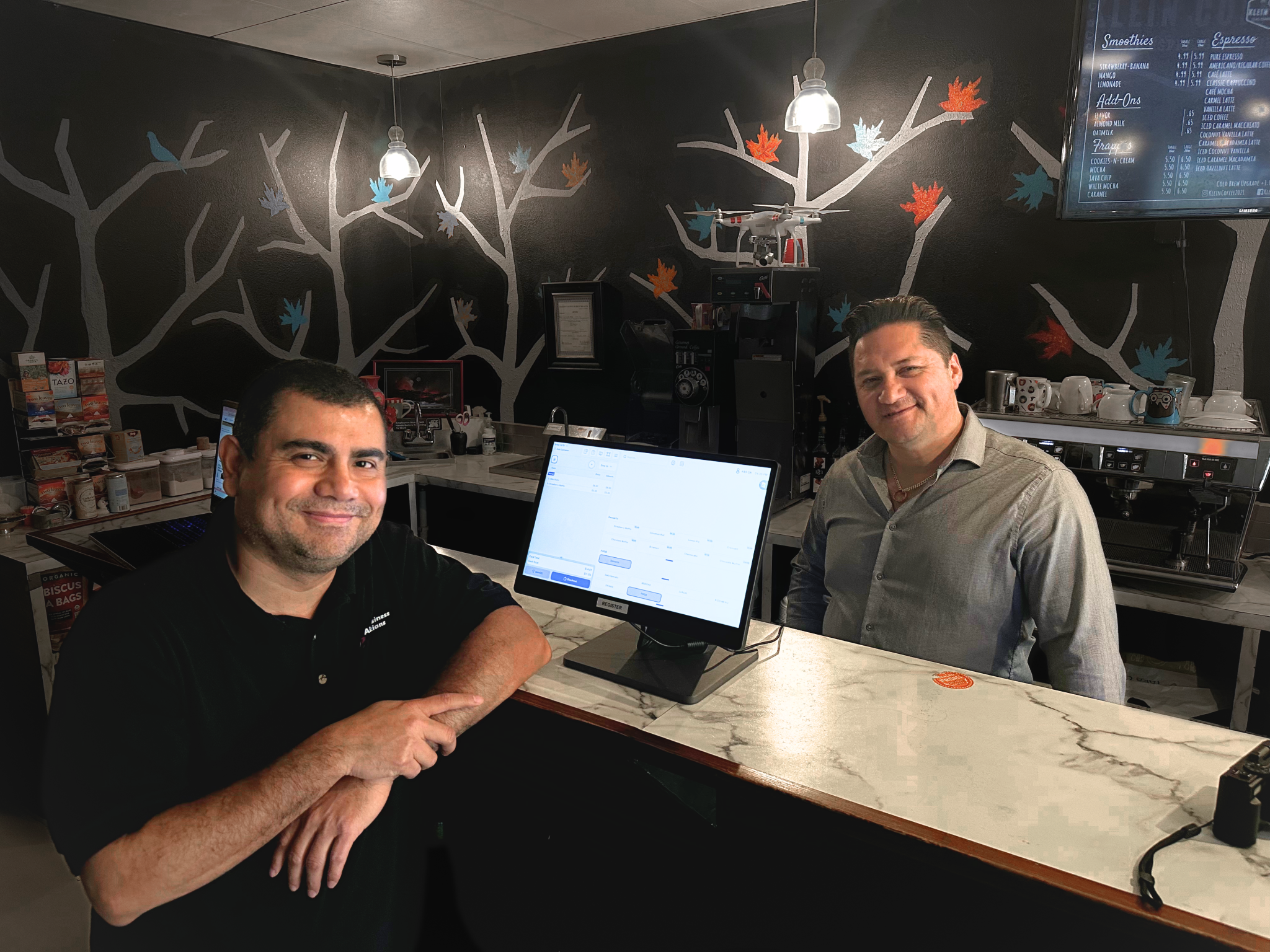 Two men smiling behind the counter of a restaurant, standing beside a touchscreen POS system on a marble countertop with a mural of abstract tree branches in the background.